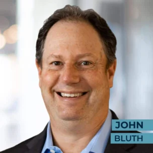 Smiling man in a suit and blue shirt with blurred background and the name "John Bluth" on the image.